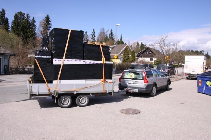 En bil drar en släpvagn lastad med staplade och inplastade dorocellblock, säkrade med spännband.  Scenen utspelar sig på en lugn gata med hus och träd i bakgrunden.