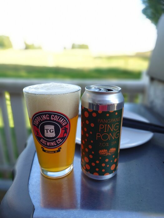 Ett glas öl från Toppling Goliath Brewing Co. och en burk PangPang Ping Pong 7.0% IPA står på ett bord utomhus med en tallrik i bakgrunden.