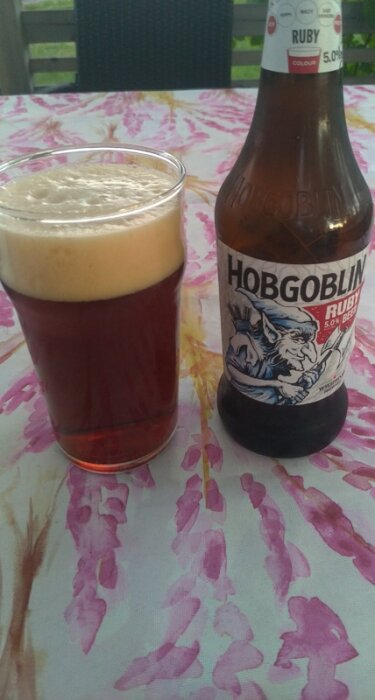 En flaska Hobgoblin Ruby öl och ett glas med skummande öl står på ett bord med en blommig duk på en altan.