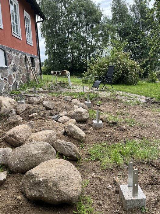 Byggarbetsplats med solstol, många stenar och en plint med metallfästen i förgrunden. Ett gammalt hus syns till vänster och ett arbetsbord står i bakgrunden.