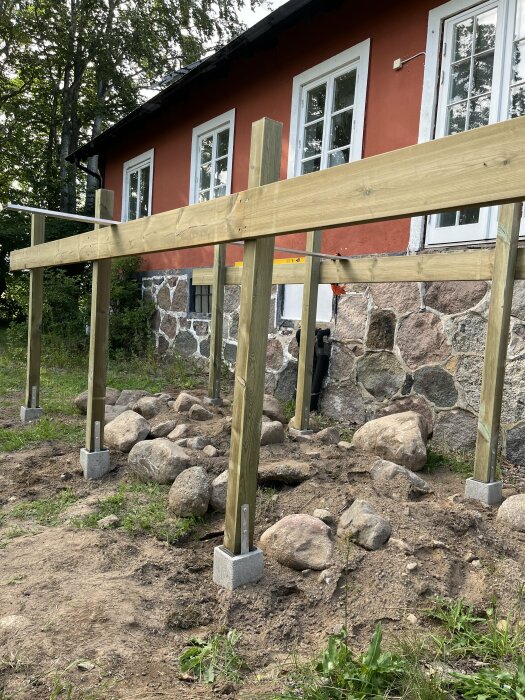 Träkonstruktion av blivande balkong med stolpar och bärlinor, monterade på betongplintar framför ett hus med stenvägg och rödmålad fasad.