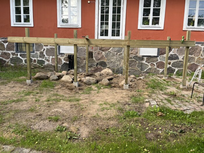 Trästolpar och balkar monterade på en grund bredvid ett stenhus, projekt för att bygga eller förstärka en balkong. Dukar och verktyg ses på marken.