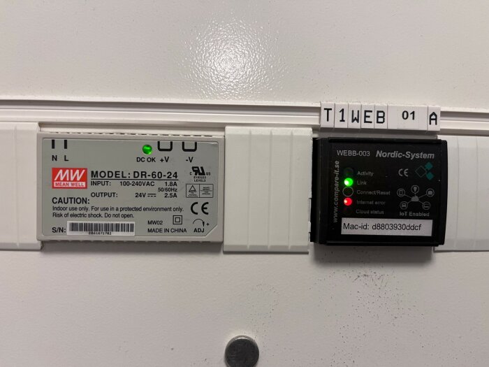 Bild på en Mean Well strömkälla och en Nordic-System enhet monterad på en vägg med en etikett som säger "T1WEB 01 A".