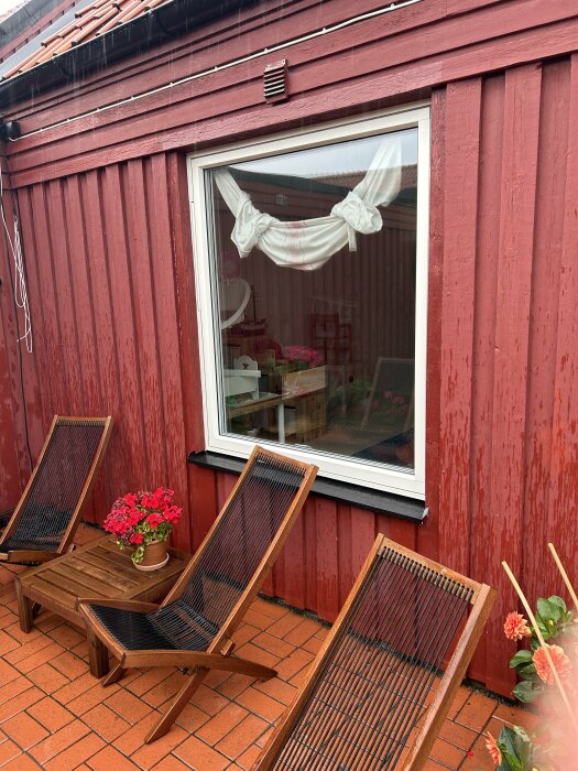 Rött trähus med ett stort fönster utan fönsterbleck. Framför huset finns tre stolar, ett bord och en kruka med röda blommor på en terrakottafärgad uteplats.