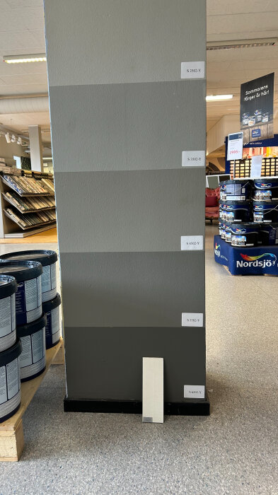 Olika grå färgnyanser (S 2502-Y, S 3502-Y, S 4502-Y, S 5502-Y, S 6502-Y) uppmålade på en pelare i en Nordsjöfärgs butik, burkar med färg syns också i bakgrunden.