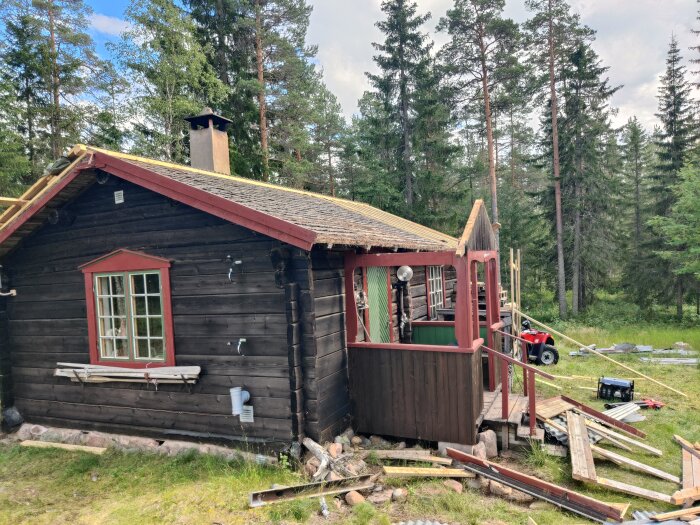 Fritidshus under takrenovering med ströläkt och bärläkt på taket, omgivet av en skog. Verktyg och byggmaterial är utspridda runtom huset.