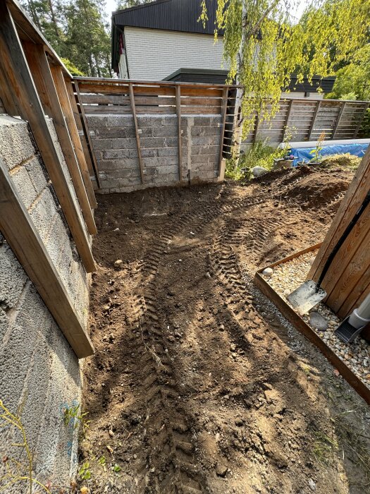 Uppgrävd yta förbered för att gjuta en betongplatta med synliga traktorspår. Bakgrunden visar ett staket, träd och ett hus.