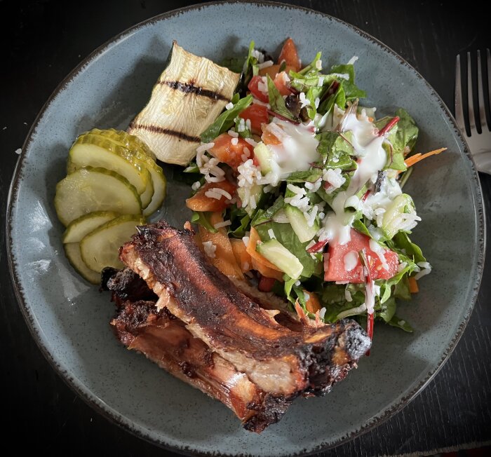 Grillade kamben, rissallad med dressing, inlagd gurka och en bit grillad squash på en tallrik.