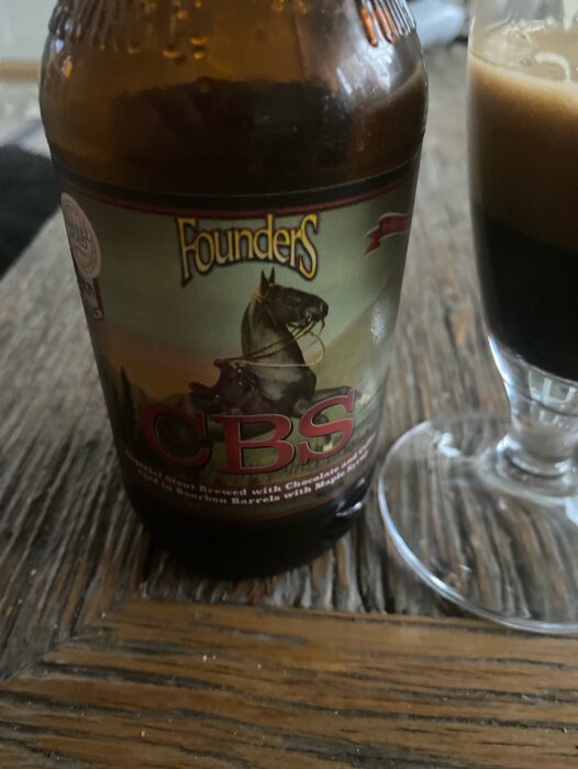 Flaska med etiketten Founders CBS, en stout med choklad och kaffe, ställd på ett bord bredvid ett glas fyllt med svart dryck och skum.