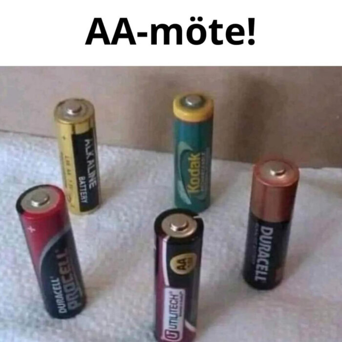 Flera AA-batterier av olika märken står upprätt på en yta med texten "AA-möte!" ovanför dem, vilket är en ordlek.