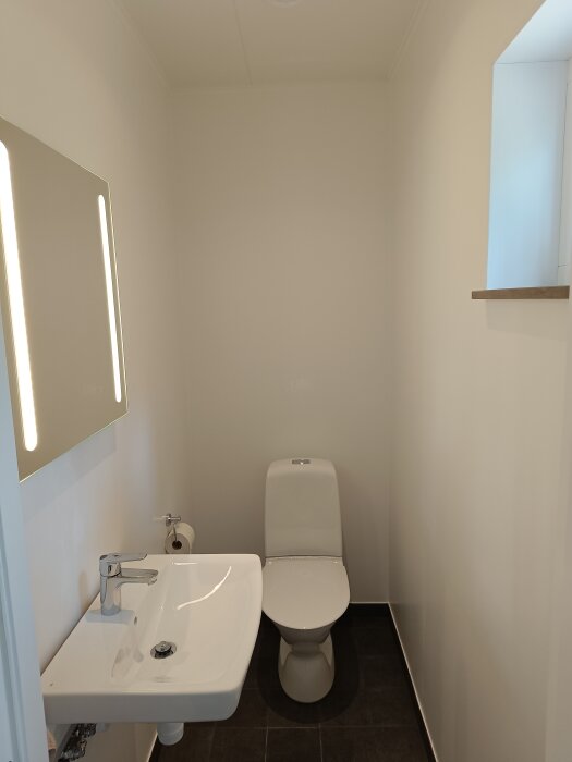 Modernt badrum med vit toalettstol och vitt handfat med spegel ovanför. Väggarna är vita och golvet är mörkt.