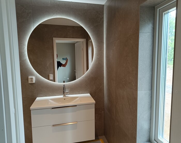 Modernt badrum med ett runt, upplyst spegel ovanför en tvättställskommod, väggar klädda i beige kakel och ett fönster till höger.