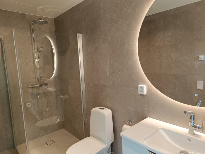 Ett modernt badrum med duschhörna, toalett, handfat och en stor rund spegel med bakgrundsbelysning, samt väggar klädda i grå kakel.