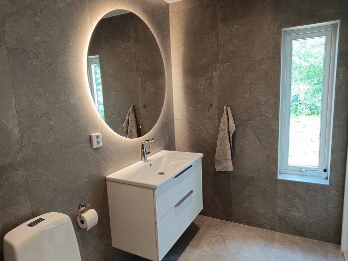 Modernt badrum med grå kakel, rund spegel med bakgrundsbelysning, vitt handfat med vit kommod, toalett, handduk på vägg och fönster med utsikt mot träd.
