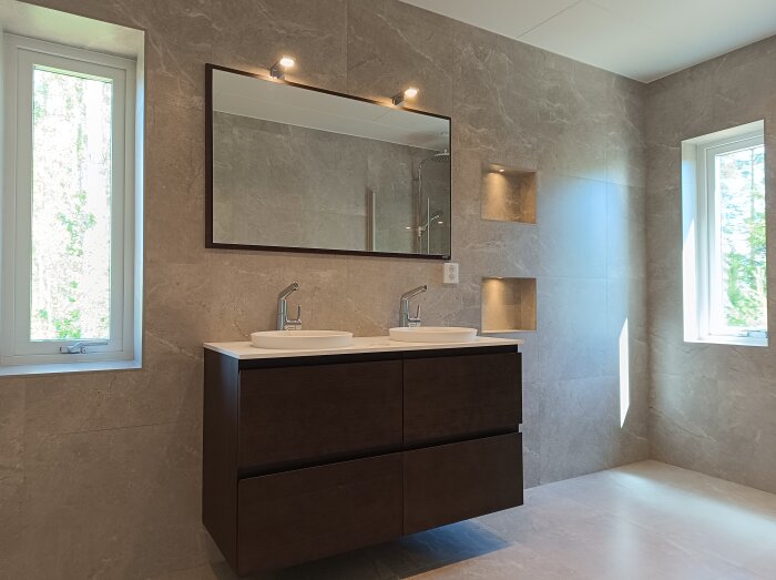 Modernt badrum med dubbla handfat, stor spegel, dusch, inbyggda hyllor och naturligt ljusinsläpp från två fönster.