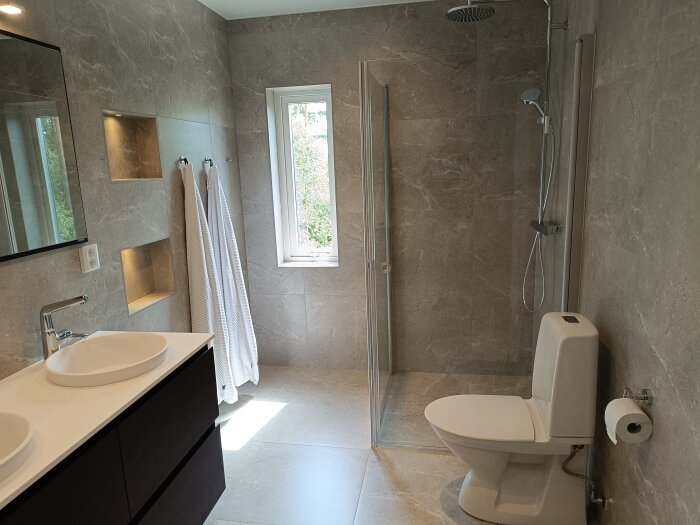 Modernt badrum med gråa kakelväggar, två handfat, en dusch med glasdörr, en toalett och handdukar hängande på väggen.