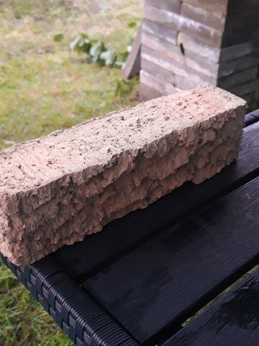 Närbild av en tegelsten med en ojämn yta, placerad på ett svart bord. Bakgrunden är en gräsyta med några gröna blad.