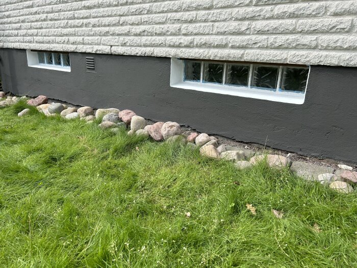 Målad husgrund med mörk färg S7502-y, omgiven av stenbelagd kant och gräsmatta, med gråvägg ovanför och små källarfönster.
