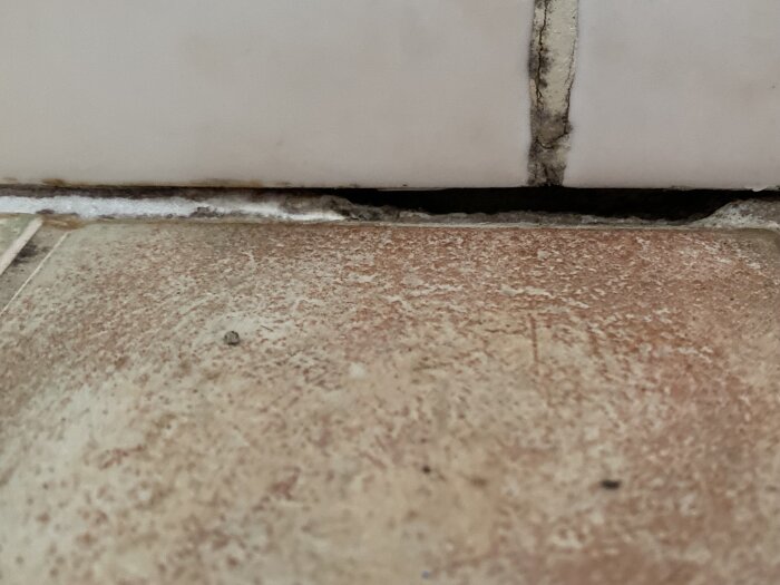 Tätningsfog i dusch med smuts och fukt mellan kakelplattor och golv, del av mjukfog borttagen, ojämna mellanrum och synliga gap bakom fogen.