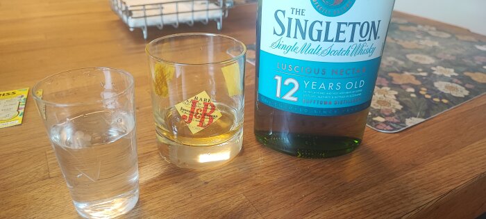 En flaska Singleton 12-årig single malt whisky på ett bord bredvid ett glas med whisky och ett glas med vatten.