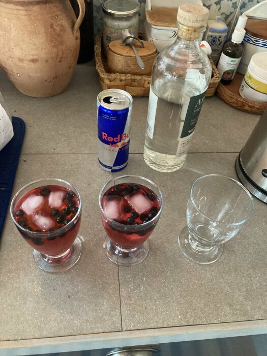 Två glas med isbitar och blåbär, en tomt glas, en Red Bull-burk och en flaska sprit står på en köksbänk.