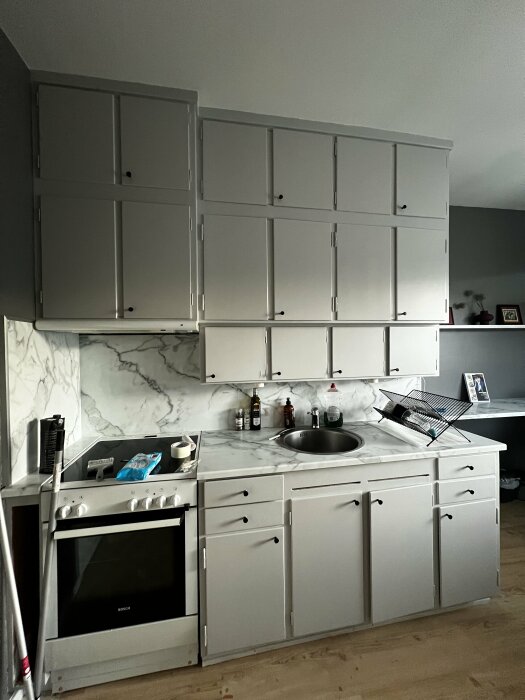 Nyrenoverat kök med nymålade skåp och väggar i ljusgrått. Vit Bosch-spis, ho och marmorlika bänkskivor. Diskställ på bänken och några flaskor på väggen.