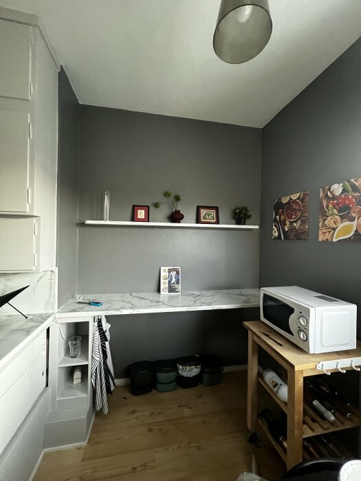 Nymålat kök med grå väggar, vit skiva, hylla med dekor, mikrovågsugn på träbord, och köksföremål på hyllor och golv.