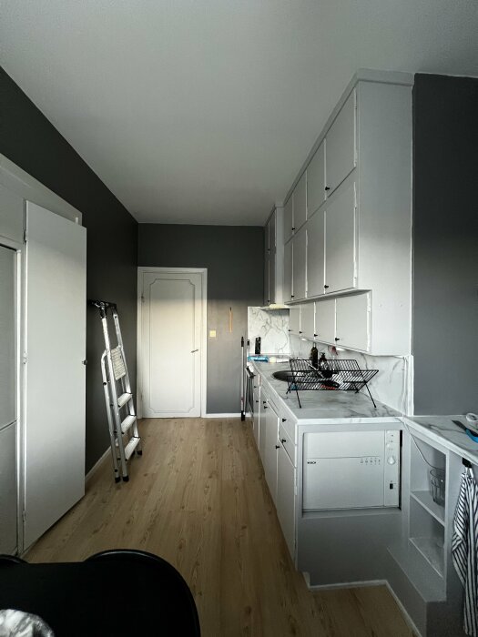 Nyrenoverat kök med vita skåpstommar och grå väggar, diskmaskin och diskställ på diskbänken, stege lutad mot väggen.