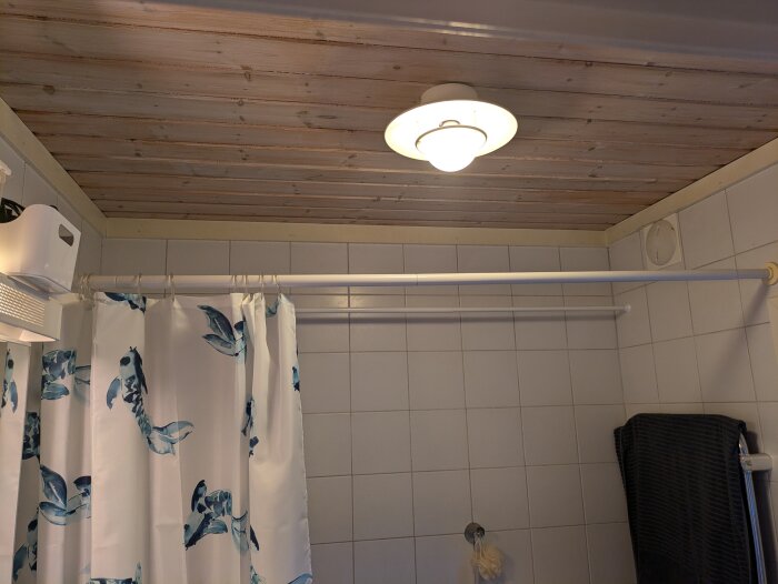 Plafondlampa i ett litet badrum med vit kakelväggar och duschdraperi med blå dekoration på en vit duschstång. Taket är av träpanel.