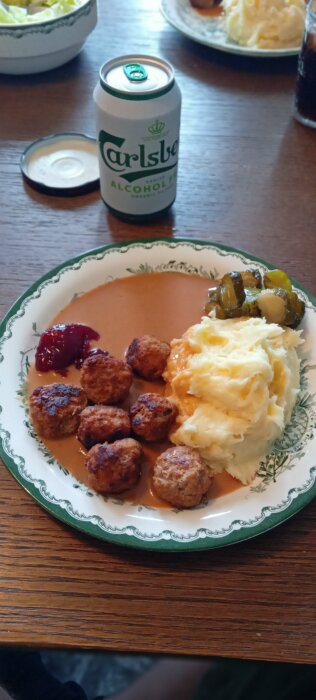 En tallrik med köttbullar, potatismos, brunsås, inlagd gurka och lingonsylt bredvid en burk alkoholfri Carlsberg öl.