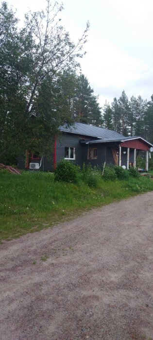 Grått hus med plåttak och röda fönsterkarmar, beläget på en grusväg med grönt gräs och träd i bakgrunden i en by utanför Kalix.