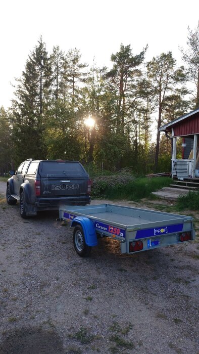 En svart Isuzu-pickup med en släpvagn märkt "Caravan 1250 XL" står parkerad på en grusad uppfart bredvid ett rött hus. Solen skiner genom träden i bakgrunden.