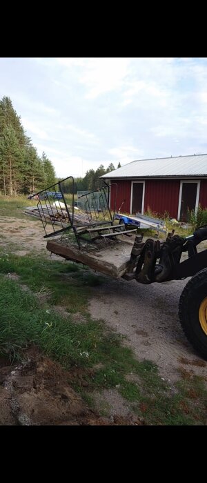 En gul grävmaskin med en fästskopa lyfter en gamal, skadad metallgrind och betongfundament. I bakgrunden syns en röd byggnad och skog.