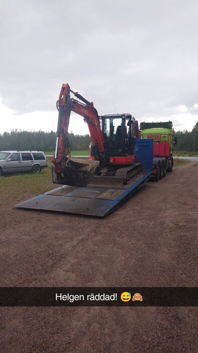 Grävmaskin av märket Kubota på en blå lastbilstrailer med grön lastbil i bakgrunden på en grusväg under molnig himmel.