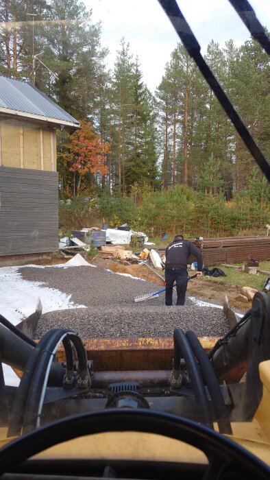 En person jämnar ut grus med en kratta bredvid ett hus under byggnation, omgiven av verktyg och skog i bakgrunden, sett från hytten på en grävmaskin.