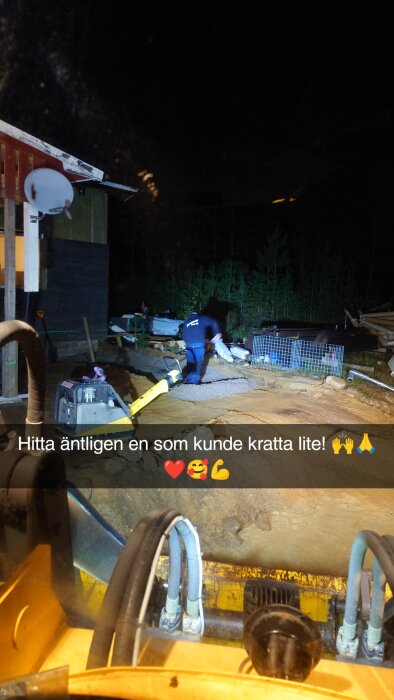 En person krattar underlag för en altan i mörkret med hjälp av strålkastare, omgiven av byggmaterial och gabioner, nära ett hus.