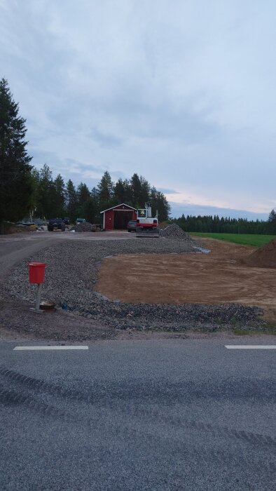 Arbetsplats för altanbygge med utlagd grus och jord, grävmaskin i bakgrunden bredvid ett rött garage, omgivande träd och fält.