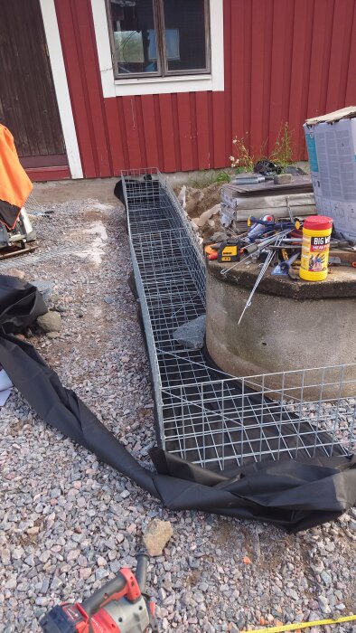 Gabioner i metallnätskorgar för ett altanprojekt, omgivna av verktyg och byggmaterial framför ett rött hus med ett fönster och en dörr.