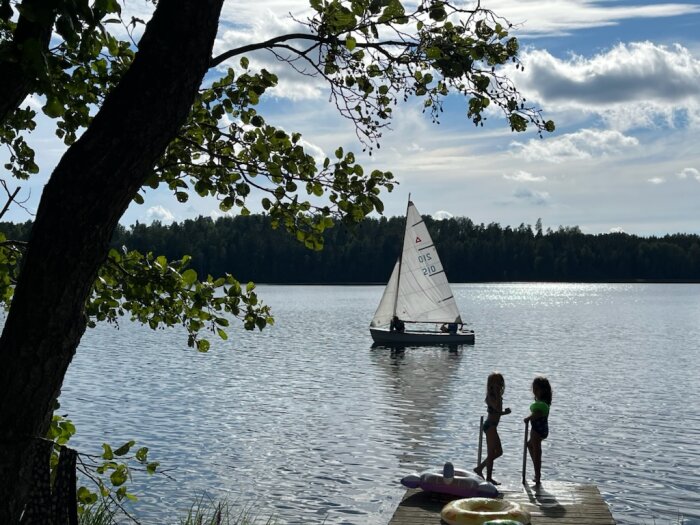 Två barn står på en brygga vid en sjö, där en segelbåt med vita segel seglar förbi. I förgrunden syns en uppblåsbar simring formad som en ananas.