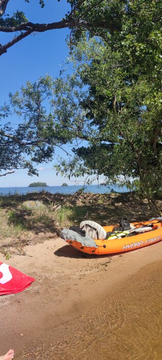 Orange uppblåsbar båt med texten "HYDRO-FORCE" ligger på en sandstrand, omgiven av grönska och med vatten och små öar i bakgrunden.