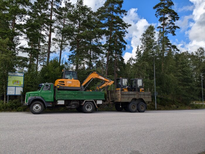 En grön lastbil med en orange grävmaskin på flaket och en trailer med två mindre grävmaskiner, parkerad på en skogsväg.