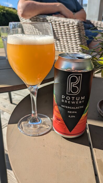 Glas med orangefärgad öl bredvid en ölburk från Potum Brewery med etiketten "Intergalactic NEIPA 6,5%" på ett bord, med en person sittandes i bakgrunden.