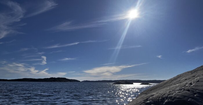Solig dag vid havet med blå himmel, vita moln och solens strålar som reflekteras i vattnet, stenar syns till höger i bild.