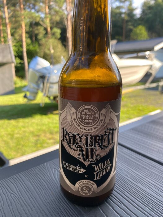 Ölflaska på ett bord utomhus med etiketten "Rye Brett Ale" och en båt i bakgrunden.