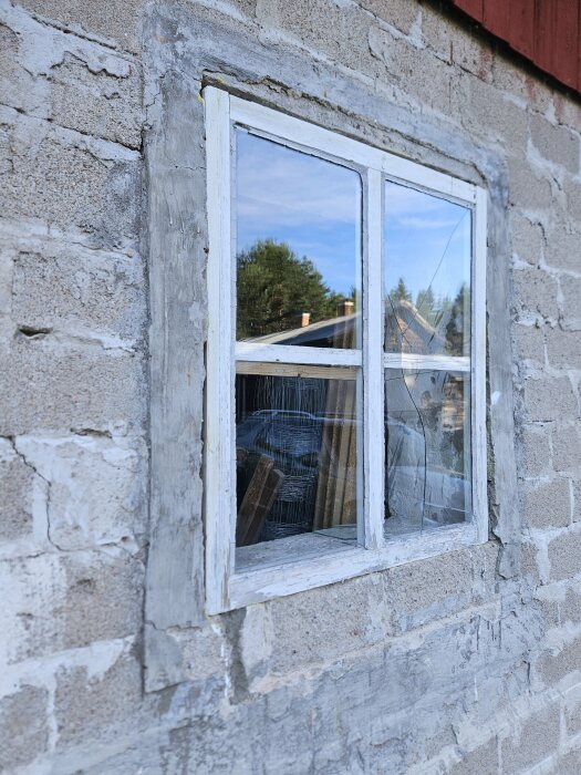 Ett gammalt fönster med skadad karm och sprucken glasruta, omgivet av omålad och opolerad murbruk på en byggnadsmur.