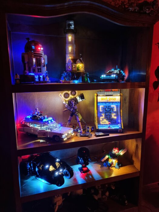 Belyst legoset med flera byggda figurer på en bokhylla, inklusive R2-D2, fyrtorn, robot, Pac-Man-maskin, Ghostbusters-bil och andra modeller.