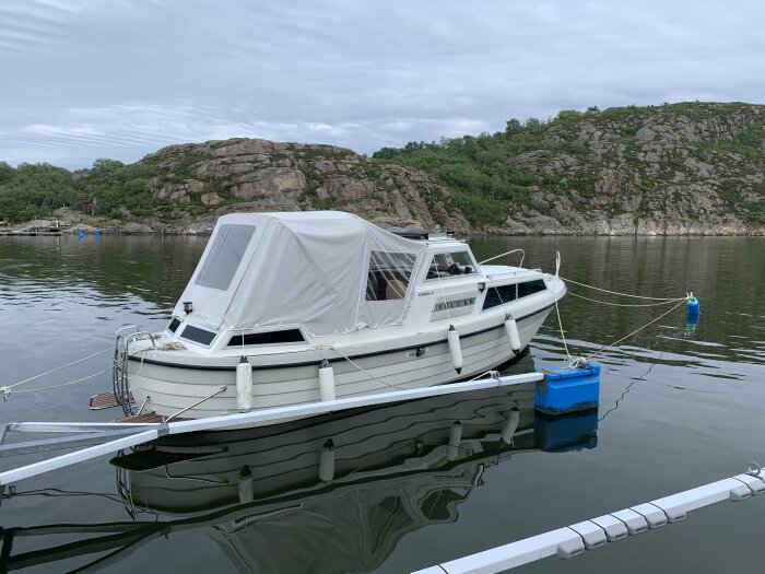 Vit båt sjösatt i lugn vik med klippor i bakgrunden. Båten är förtöjd till piren med rep och bakgrund av grönska och låga klippformationer.