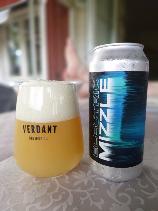 Ölglas märkta "Verdant Brewing Co." och en burk "Electric Mizzle" IPA står på ett bord. Bakom syns ett suddigt fönster och någon som vilar.