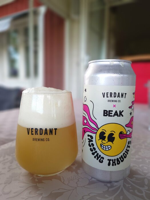 En ölglas med Verdant-logo och en ölburk med texten "Verdant x Beak, Passing Thoughts" står på ett bord.