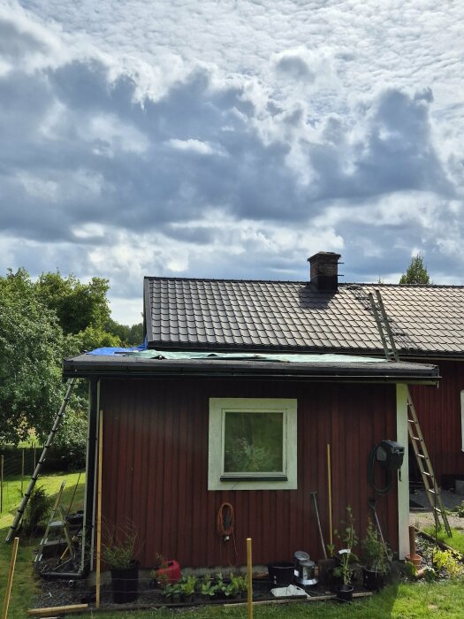 Tillbyggnad med platt tak som är täckt med en presenning, fäst vid hus med sluttande tak. Stegar stödda mot taket. Molnig himmel i bakgrunden.